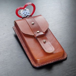 May include: A brown leather case with a flap and strap closure. A red heart-shaped charm with a Dalmatian dog figurine is attached to the top. The case is rectangular and appears to be handmade.