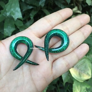 K&ouml;nnte beinhalten: Ein Paar gr&uuml;ne Glitzer-Ohrmanschetten, jede in Form eines gekr&uuml;mmten, offenen Rings. Die Manschetten werden auf einer Hand gezeigt, wobei der gr&uuml;ne Glitzer das Licht reflektiert.