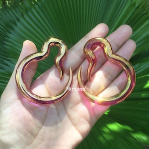 May include: Two gold and pink heart-shaped glass plugs for stretched ears. The plugs are shown on a hand.