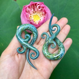 May include: A pair of green and blue glass ear gauges with a swirling design. The gauges are made of dichroic glass, which means they change colour depending on the angle of light.