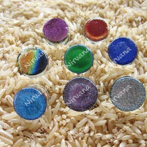 May include: Eight round glass cabochons with iridescent and solid colors. The colors include purple, red, green, blue, and silver. The cabochons are arranged in a circle on a bed of brown rice.