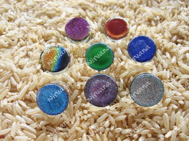 May include: Eight round glass cabochons with iridescent and solid colors. The colors include purple, red, green, blue, and silver. The cabochons are arranged in a circle on a bed of brown rice.