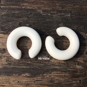 May include: Two white ceramic hoop earrings, each with a rounded, slightly curved shape. The earrings are displayed on a wooden surface.