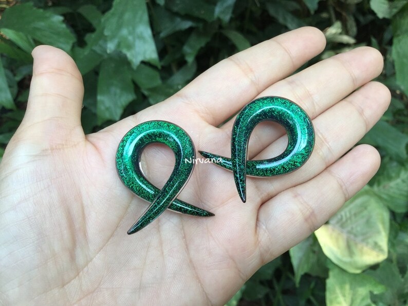 K&ouml;nnte beinhalten: Ein Paar gr&uuml;ne Glitzer-Ohrmanschetten, jede in Form eines gekr&uuml;mmten, offenen Rings. Die Manschetten werden auf einer Hand gezeigt, wobei der gr&uuml;ne Glitzer das Licht reflektiert.