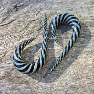 Puede incluir: Un par de dilatadores de oreja de vidrio en espiral de color negro y blanco. Los dilatadores están hechos de vidrio y tienen un diseño en espiral. Tienen un diámetro de aproximadamente 2,5 cm.