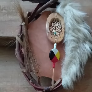 Puede incluir: Un atrapasueños de cuero marrón con una pluma blanca, una cuenta de madera con un diseño colorido y dos plumas marrones que cuelgan de la parte inferior.