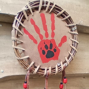 Peut inclure: Un attrape-rêves en cuir brun avec une empreinte de main rouge et une empreinte de patte noire au centre. L'attrape-rêves est orné de plumes noires et de perles rouges.