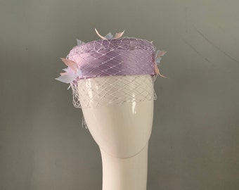 Bonnet carré lilas orné d'une bande et d'un noeud en satin de soie voilant Merry Widow, rehaussés de plumes bleu pâle et roses.