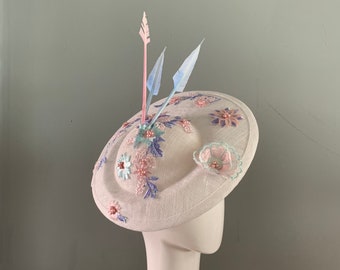 White saucer hat fascinator adorned with pastel Pink and Blue lace appliques finished with dramatic arrowhead feathers.