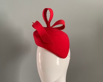 May include: A bright red felt hat with a large bow on top. The hat is perched on a white mannequin head.