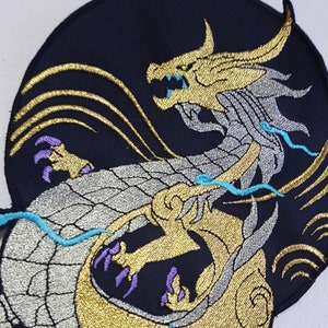Könnte beinhalten: Gestickter Bügel-Patch mit einem silbernen und goldenen Drachen mit türkisfarbenen und violetten Akzenten. Der Drache ist zu einem Kreis zusammengerollt, mit dem Kopf nach vorne gerichtet.