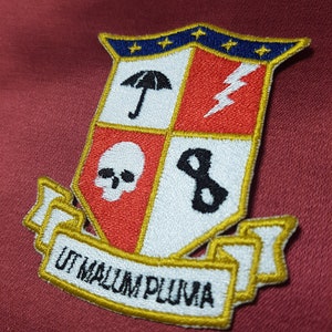 May include: A gold embroidered patch with a shield design. The shield is divided into four sections, each with a different symbol. The top left section has an umbrella, the top right section has a lightning bolt, the bottom left section has a skull, and the bottom right section has a gear. The shield is surrounded by a gold border and a banner with the text "UT MALLUM PLUVIA".