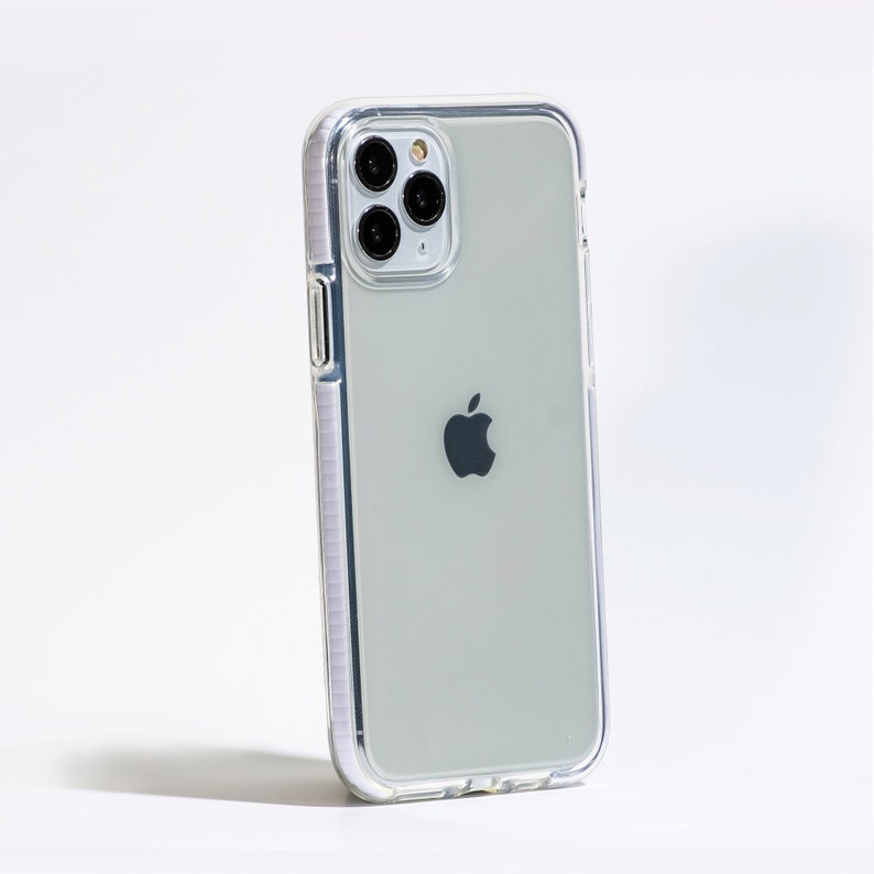 May include: A clear phone case with a white border, protecting a silver iPhone with a black Apple logo. The phone has a triple lens camera system.