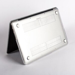 May include: A clear, transparent protective hard shell case attached to the underside of a silver laptop. The case features four circular rubber feet at the corners and integrated ventilation slats, with the text MacBook Pro visible.