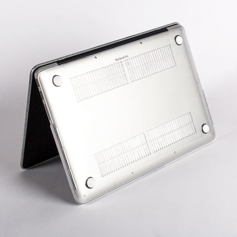 May include: A clear, transparent protective hard shell case attached to the underside of a silver laptop. The case features four circular rubber feet at the corners and integrated ventilation slats, with the text MacBook Pro visible.