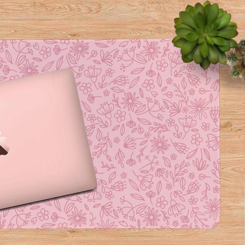 K&ouml;nnte beinhalten: Ein pinkfarbener Laptop liegt auf einer passenden pinkfarbenen Schreibtischunterlage. Die Unterlage hat ein zartes Blumenmuster in einem helleren Rosaton. Daneben steht eine gr&uuml;ne Sukkulentenpflanze auf einer Holzoberfl&auml;che.