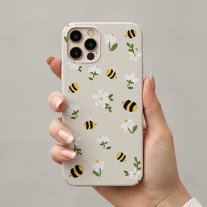 Könnte beinhalten: Eine hellbeige Smartphone-Hülle mit einem wiederkehrenden Muster aus kleinen gelb-schwarzen Hummeln, durchsetzt mit weißen gänseblümchenartigen Blüten mit grünen Stielen und Blättern. Die Hülle hat eine Aussparung für ein Dreifach-Kamerasystem.