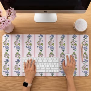 Puede incluir: Una alfombrilla de escritorio rectangular con un patrón vertical repetitivo de rayas rosa claro intercaladas con columnas florales de flores azules y rosas, hojas verdes y pequeñas mariposas azules sobre un fondo rosa pálido.