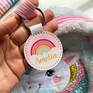 Peut inclure: Un porte-clés rond avec un arc-en-ciel et le nom "Amelia" en orange. Il est attaché à une sangle rose et un anneau argenté. En arrière-plan, un sac en peluche sur le thème de la licorne avec une fermeture éclair rose.