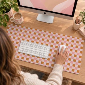 May include: A rectangular desk mat featuring a repeating checkered pattern in alternating shades of soft pink and mustard yellow. The mat is positioned on a wooden surface beneath a white wireless keyboard and a white computer mouse.