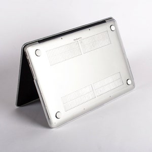 May include: A clear, transparent protective hard shell case attached to the underside of a silver laptop. The case features four circular rubber feet at the corners, ventilation slats, and the text MacBook Pro printed on the center edge.