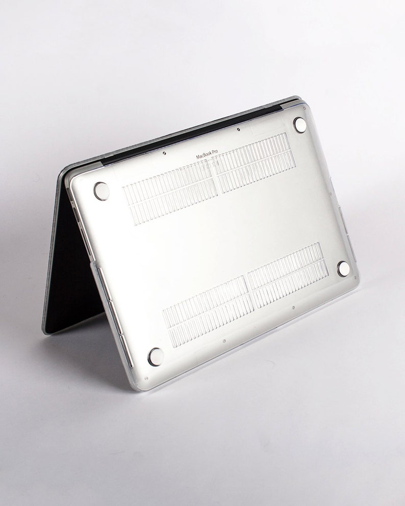 May include: A clear, transparent protective hard shell case attached to the underside of a silver laptop. The case features four circular rubber feet at the corners, ventilation slats, and the text MacBook Pro printed on the center edge.