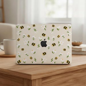 Könnte beinhalten: Beige Laptop-Hülle mit einem Muster aus gelb-schwarzen Bienen, weißen Blumen und grünen Blättern. Die Hülle hat ein schwarzes Apple-Logo in der Mitte. Das Design ist skurril und von der Natur inspiriert.