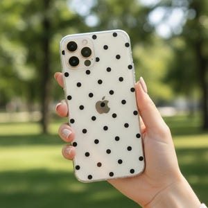 May include: Clear phone case with a black polka dot pattern. The case is held in a hand, and the Apple logo is visible on the back. The phone has a multi-lens camera.