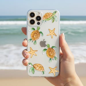 May include: Clear phone case featuring a watercolor design of sea turtles and starfish. The turtles have brown shells and green limbs, while the starfish are yellow. The case is held against a backdrop of a beach and ocean.