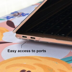 May include: A rose gold laptop encased in a clear protective shell, showing the side profile with two USB-C ports. A thin black line points to the ports with the text Easy access to ports printed below the laptop on a floral patterned surface.