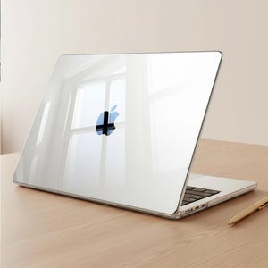 Op de afbeelding: Een transparante laptophoes met het Apple-logo. De laptop is open en toont het toetsenbord. De hoes ligt op een houten oppervlak met een pen. De hoes reflecteert het licht van een raam.