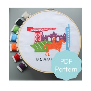 May include: A white embroidery hoop with a cross-stitch design of the Glasgow skyline. The design includes a crane, buildings, a horse, and the word "GLASGOW". The hoop is surrounded by colourful embroidery floss.