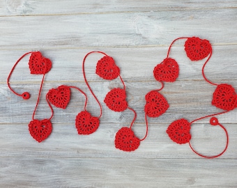 May include: A red crocheted heart garland with 10 hearts strung on a white string. The garland is laid out on a light gray wooden surface.