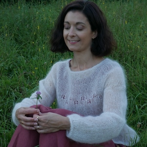 Peut inclure: Une femme portant un pull blanc en tricot ajouré est assise dans un champ d'herbe verte, tenant une petite fleur violette.
