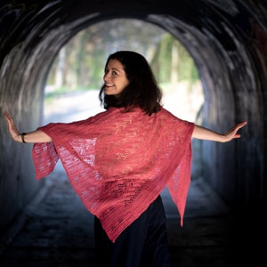 May include: A woman wearing a pink crocheted shawl stands in a tunnel with graffiti on the walls. She is smiling and looking over her shoulder.