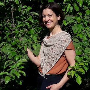 May include: A woman wearing a light brown top and black pants stands in front of a green leafy background. She is wearing a beige crocheted shawl with a lacy pattern.