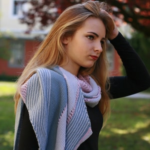 Peut inclure: Une écharpe tricotée dans les tons de bleu, gris et rose. L'écharpe est drapée autour du cou et des épaules. Le motif présente un motif rayé avec une bordure délicate en tissage ajouré. L'écharpe est en fil doux et texturé.