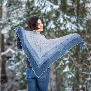 May include: A woman wearing blue jeans and a gray and blue knitted shawl with a lacy pattern. The shawl is draped over her shoulders and she is smiling at the camera.