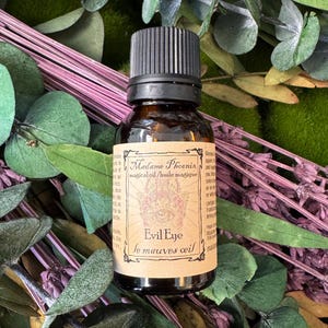 May include: A small amber glass bottle with a black screw-top cap and a tan label. The label features the text Madame Phoenix, magical oil, and Evil Eye, along with a graphic of a hamsa hand. The bottle is surrounded by dried green leaves.