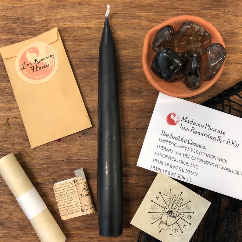 May include: A black candle, a small brown envelope with a white yin yang symbol and the text "Linx Removing Herbs Phoenix", a small brown bowl with clear and black stones, a rolled up parchment scroll, a small brown envelope with a white label and text, and a white card with black text and a black and white illustration of a hand with an eye in the palm. The text on the card reads "Madame Phoenix Jinx Removing Spell Kit This Spell Kit Contains: 1 Dipped Candle with Cotton Wick 1 Herbal Sachet of Dressing Powder for You 1 Anointing Oil Blend 1 Parchment Talisman 1 Parchment Scroll"