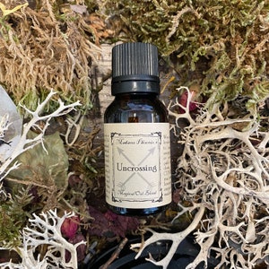 Könnte beinhalten: Eine kleine braune Glasflasche mit schwarzem Verschluss. Das Etikett lautet "Madame Phoenix's Uncrossing Magical Oil Blend". Die Flasche ist von getrocknetem Moos und anderen natürlichen Elementen umgeben.
