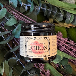 May include: A dark brown glass jar of lotion with a beige label featuring a red lip print and the text "kiss me quick LOTION embrasse-moi vite 2oz/60ml". The label includes decorative black line art borders and small printed text details.