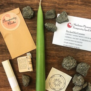 May include: A green candle, a small brown paper bag with the words "Prosperity Herbs" printed on it, and a variety of small, dark gray stones. The text "This Spell Kit Contains: 1 Dipped Candle with Cotton Wick, 1 Herbal Sachet of Dressing Powder, 1 Anointing Oil Blend, 1 Parchment Talisman, 1 Parchment Scroll" is printed on a white piece of paper.