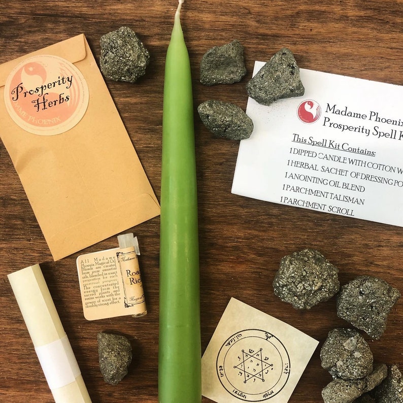 May include: A green candle, a small brown paper bag with the words "Prosperity Herbs" printed on it, and a variety of small, dark gray stones. The text "This Spell Kit Contains: 1 Dipped Candle with Cotton Wick, 1 Herbal Sachet of Dressing Powder, 1 Anointing Oil Blend, 1 Parchment Talisman, 1 Parchment Scroll" is printed on a white piece of paper.