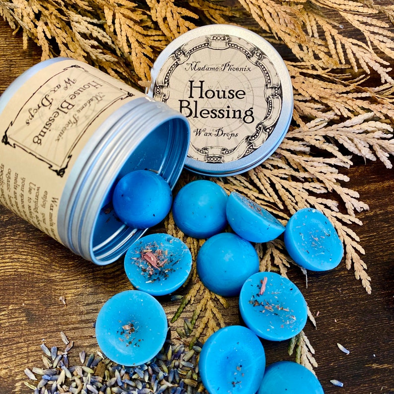 May include: A silver tin with a blue lid containing blue wax melts. The tin has a label that reads "Madame Phoenix House Blessing Wax Drops". The wax melts are scattered around the tin on a wooden surface.