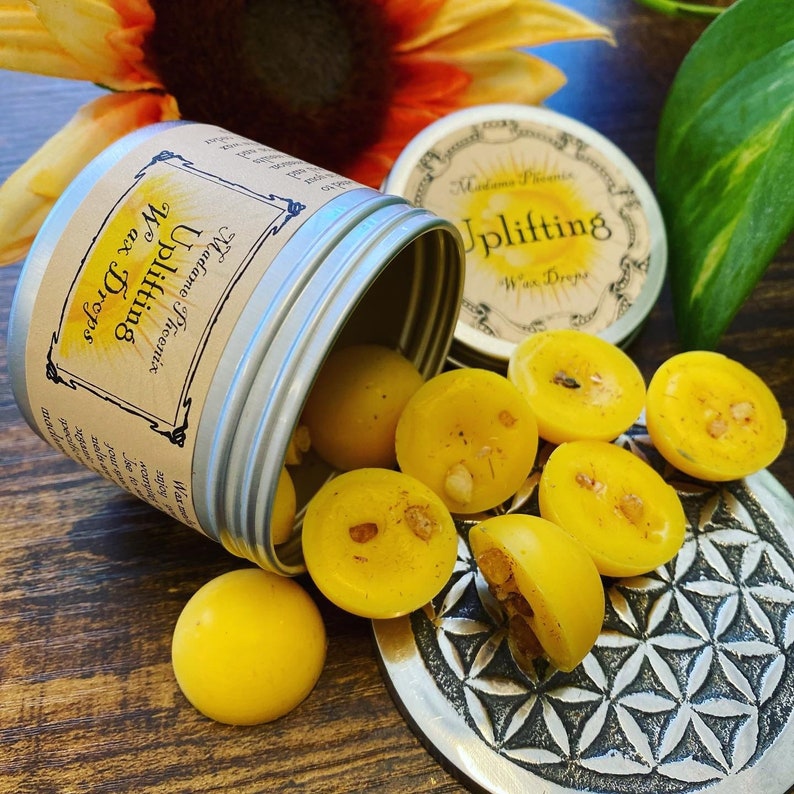 May include: A tin of yellow wax melts with the label "Uplifting Wax Drops" and the text "Madame Phoenix" on the lid. The tin is open and the wax melts are spilling out onto a silver tray with a geometric pattern.