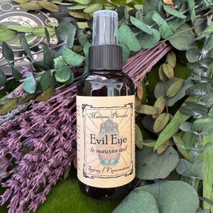 May include: A dark brown spray bottle with a black cap and a clear plastic top. The label reads "Evil Eye" and "Madame Phoenix". The bottle is surrounded by green leaves, purple lavender, and other greenery.
