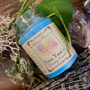 Könnte beinhalten: Ein blaues Glasgefäß mit einer Kerze und einem weißen Etikett mit der Aufschrift "Madame Phoenix's New Year's Blessing Candle". Das Etikett zeigt eine farbenfrohe Feuerwerks-Illustration.