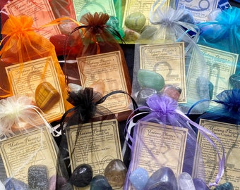 May include: A collection of twelve sheer organza drawstring pouches in various colors, each containing assorted polished gemstones and a printed card labeled Madame Phoenix's Crystal Set for different zodiac signs, arranged on a dark surface.