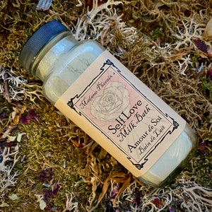May include: A glass jar filled with white powder, labelled "Self Love Milk Bath Amour de Soi Bain de Lait" with a rose illustration. The jar is sitting on a bed of dried moss and flowers.